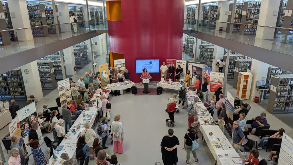 Besucherinnen und Besucher informieren sich bei Ständen und einer Rede auf der Veranstaltung „Plattform 55+“ in einer Bibliothek.